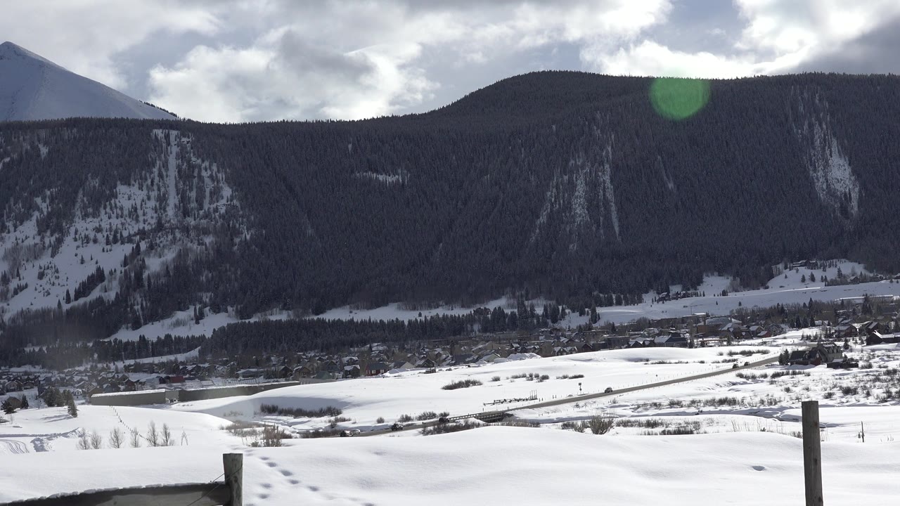 A Crested Butte, Colorado winter valley view shows snowfields, distant buildings, a dark forested ridge, foreground fence posts, cloudy light, and no visible people.