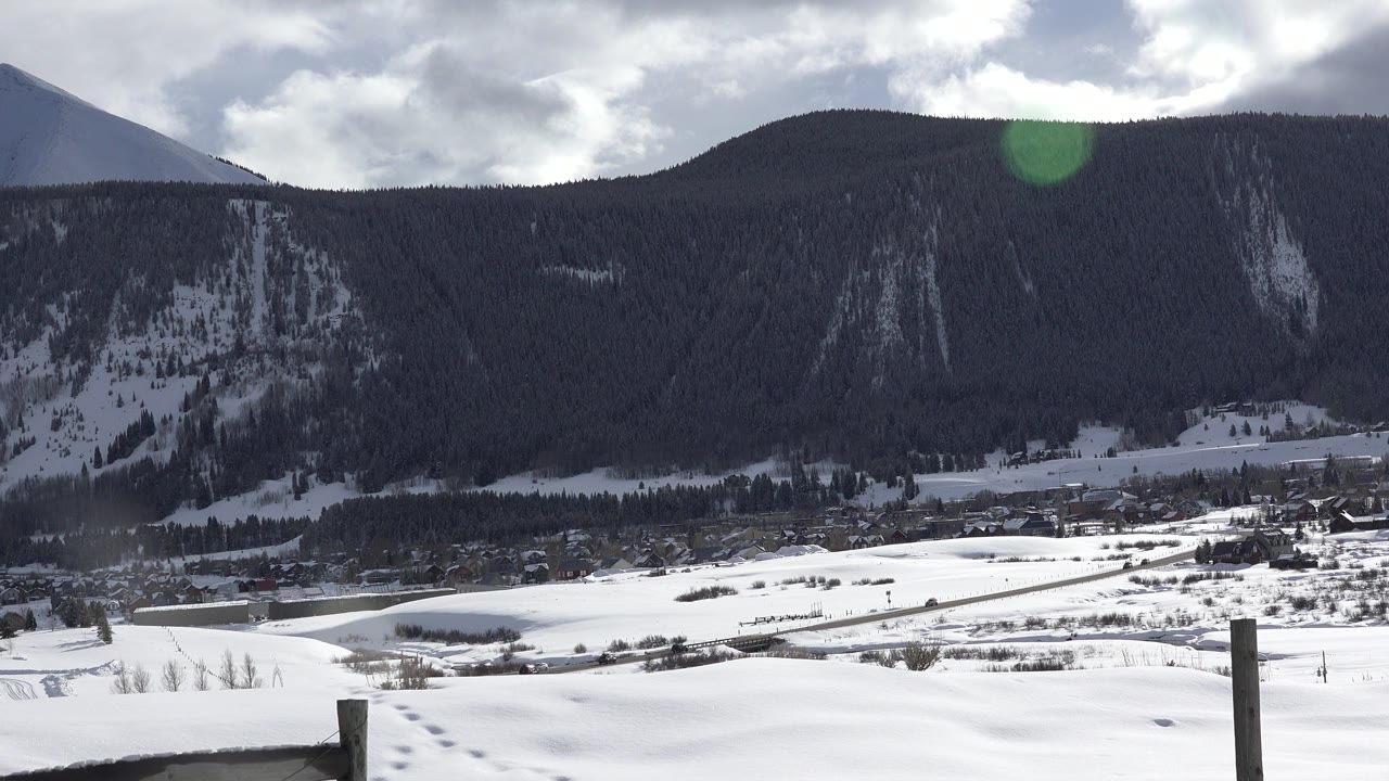 The second frame holds on the Crested Butte snowfield, with a dark wooded mountain across the background, small distant structures near the base, and no visible people.