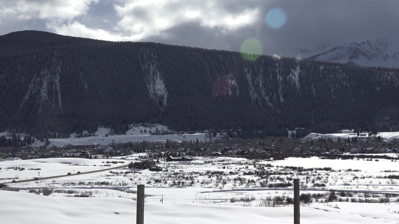Wide Crested Butte, Colorado winter landscape with snow-covered ground, distant buildings, dark forested slopes, a higher snowy ridge, lens flare, and no visible people.