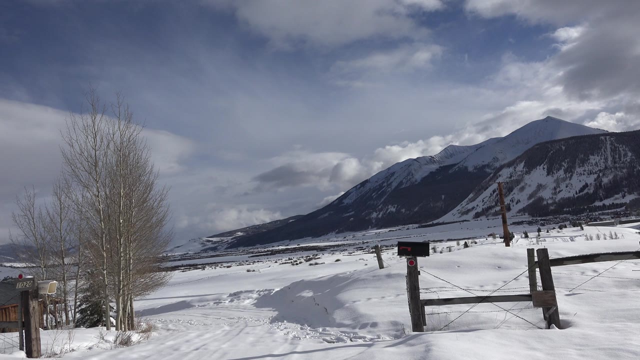 Opening frame of a snowy Crested Butte landscape with a wooden gate, leafless trees, distant mountain, broken clouds, and no visible people.