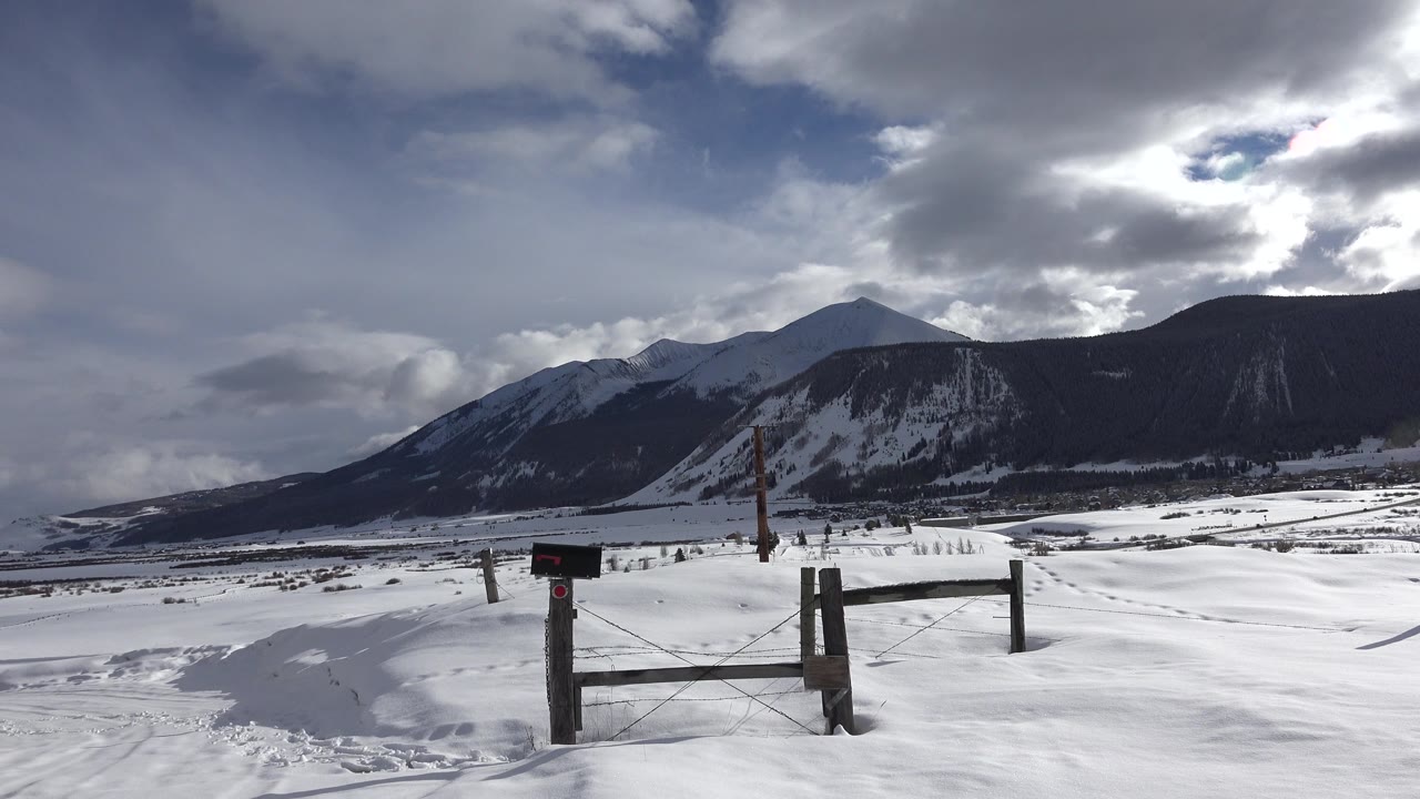 Snowy field with a wooden gate, fence posts, dark mountains, and cloudy sky.