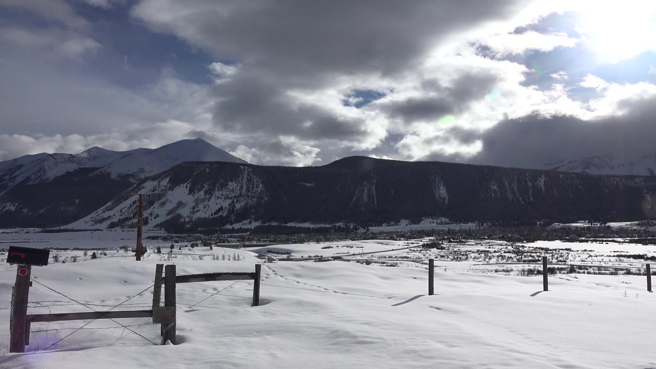 Opening frame in Crested Butte, Colorado showing a snow-covered field, wire fence, dark mountain ridge, bright cloud break, and no visible people.