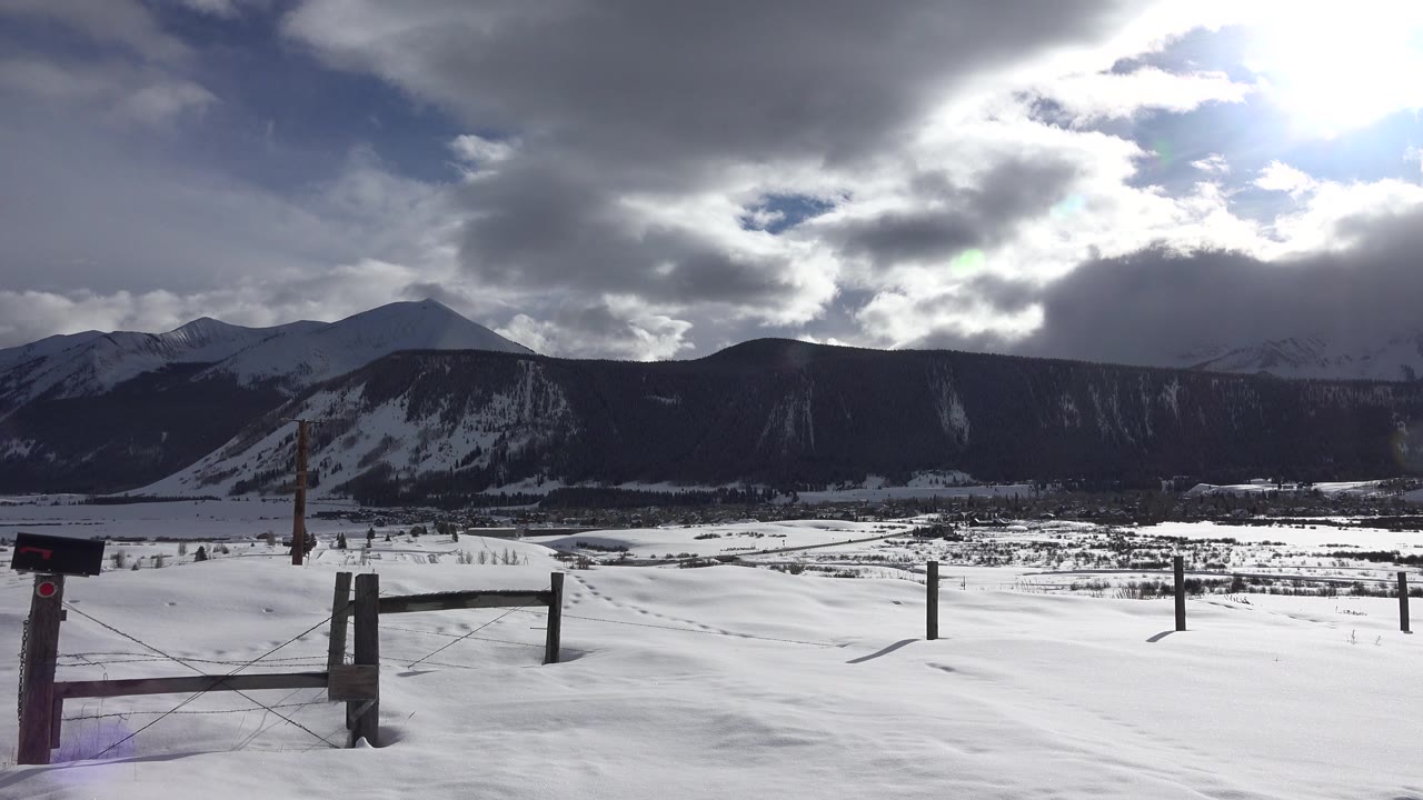 Crested Butte winter frame with a fence and roadside box in the left foreground, open snowfield, dark mountain ridge, sunlit clouds, and no visible people.