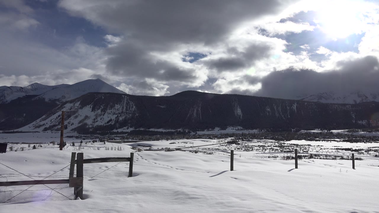 Bright winter sky over snowy valley, fence posts, and distant mountains.