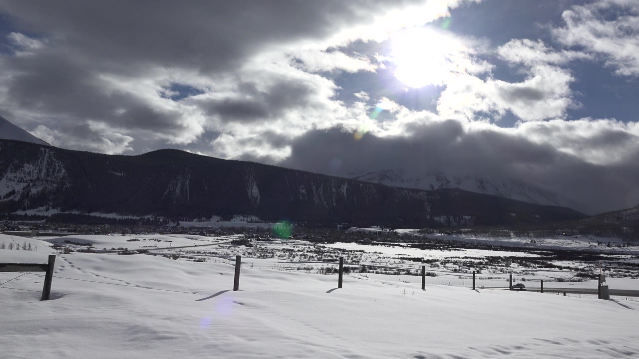 Snowy roadside with fence posts, mountains, bright sun, and heavy clouds.