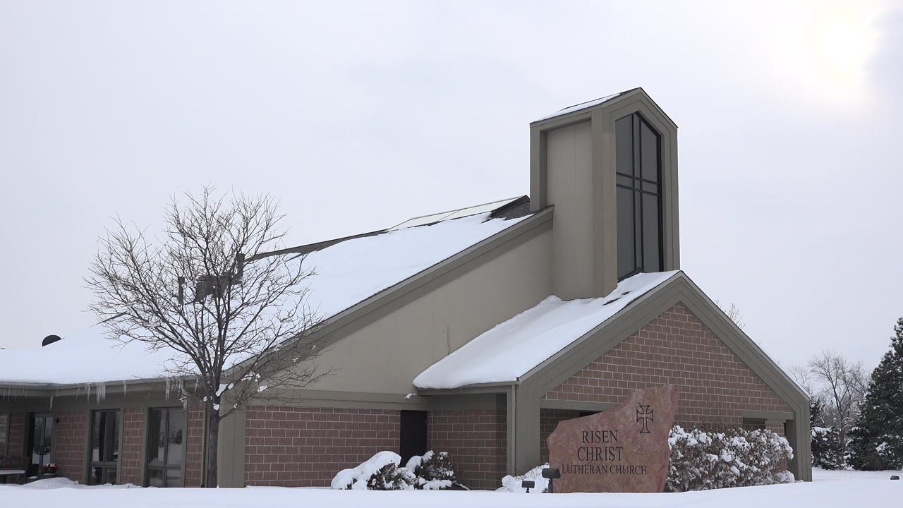 Close exterior view of a snow-covered church in Arvada, Colorado, with a brick wall, tan roofline, bare tree, stone sign, overcast sky, and no visible people.