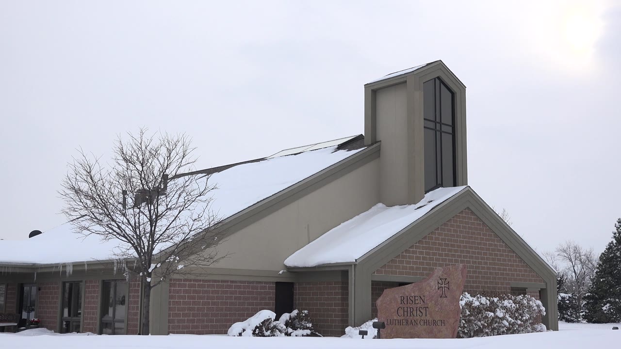 Snow blankets the church lawn and roof in Arvada, Colorado, while the brick facade, tall window tower, bare tree, and partially readable stone sign remain in view with no visible people.