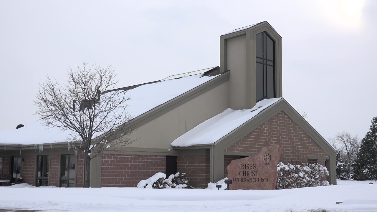 Snow-covered tan church exterior with bare tree and stone sign.