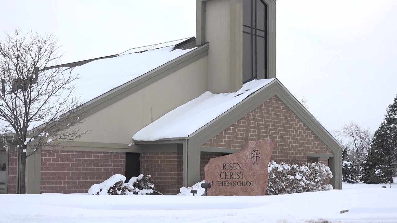 A similar Arvada church view holds on the taupe wall, brick facade, snowy roof edge, stone sign, bare tree, and empty snow-covered foreground.