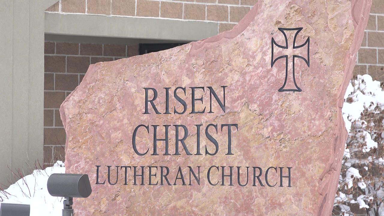 Carved Risen Christ Lutheran Church rock sign beside snow and a brick building.