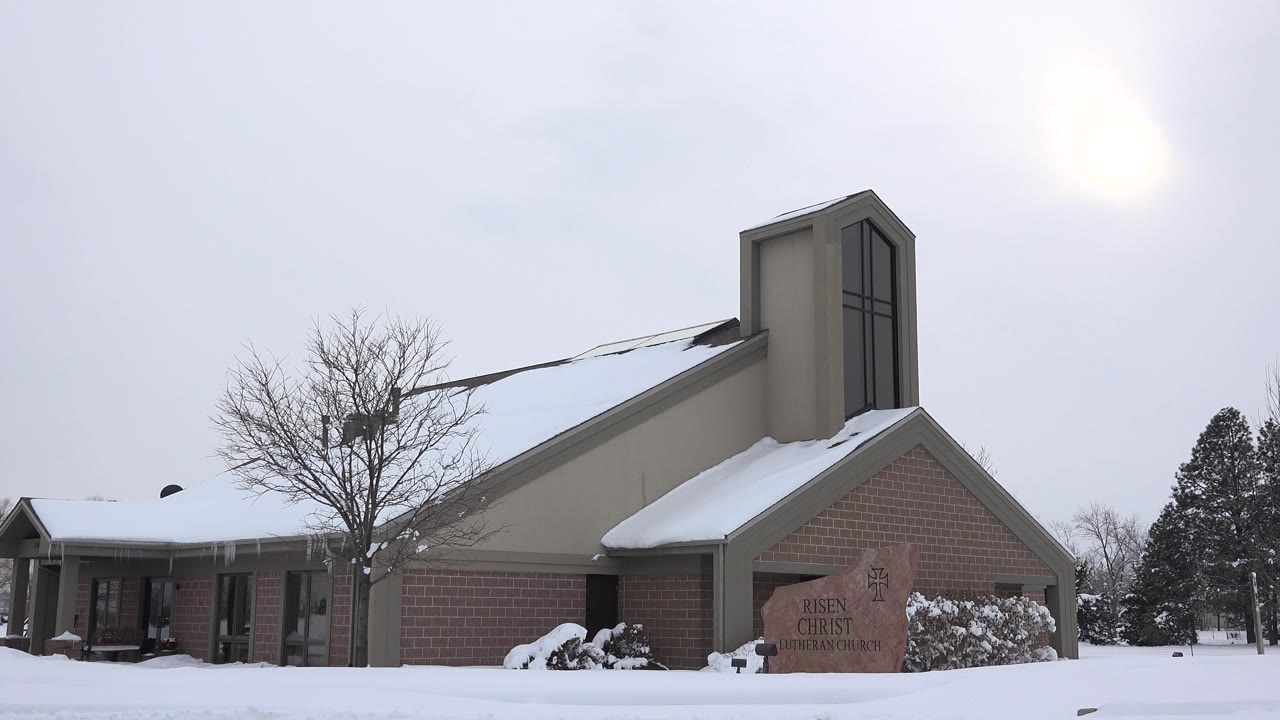 The Arvada church remains centered with a snow-covered roofline, brick side wall, stone sign near the entrance, and no visible people in the winter scene.