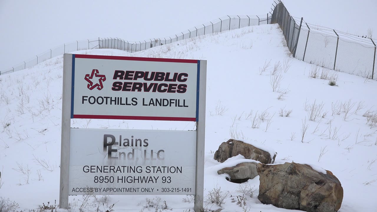A landfill entrance sign remains prominent beside large rocks, dry grass, and a snow-covered fenced slope in Arvada, Colorado, with no visible people.