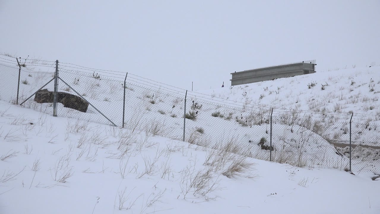 Snow-covered hillside in Arvada, Colorado with a chain-link fence, dry grasses, a small roadside structure, and no visible people at the opening moment.