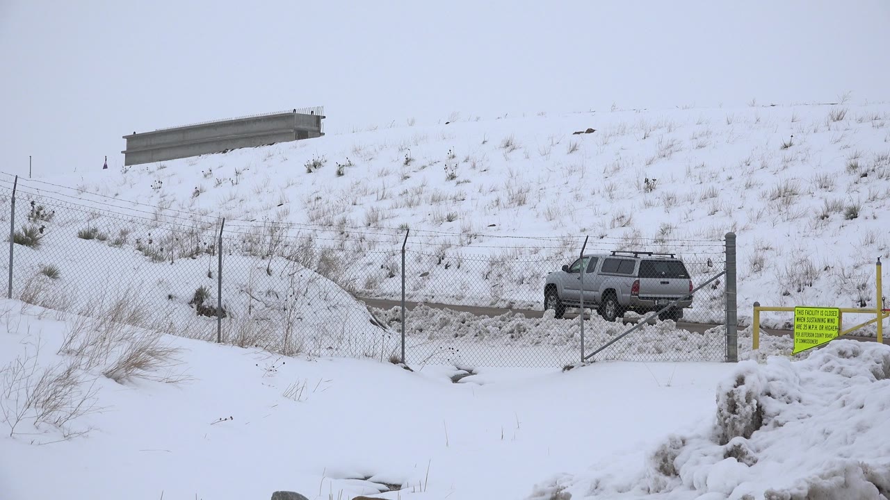 The SUV remains near the snowy roadside gate in Arvada, Colorado, framed by chain-link fencing, yellow posts, a snow pile, and no visible people.