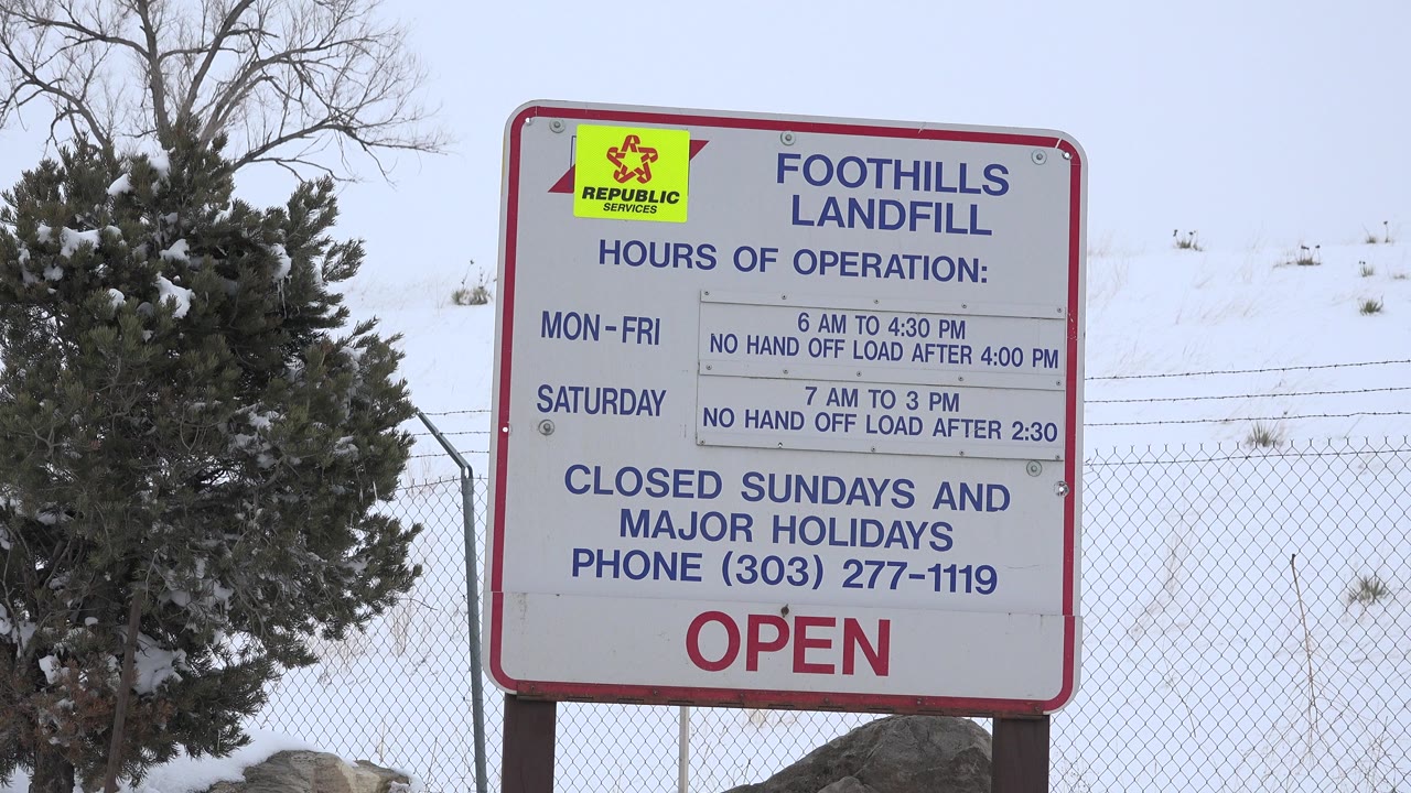 Foothills Landfill hours sign beside a snowy evergreen and fence.