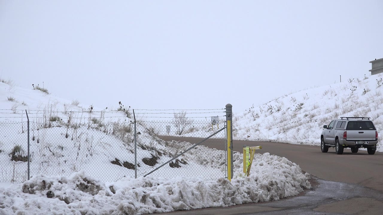 Grey SUV farther up the plowed road in Arvada, Colorado, framed by piled snow, chain-link fencing, yellow roadside posts, bare winter brush, and no visible people.