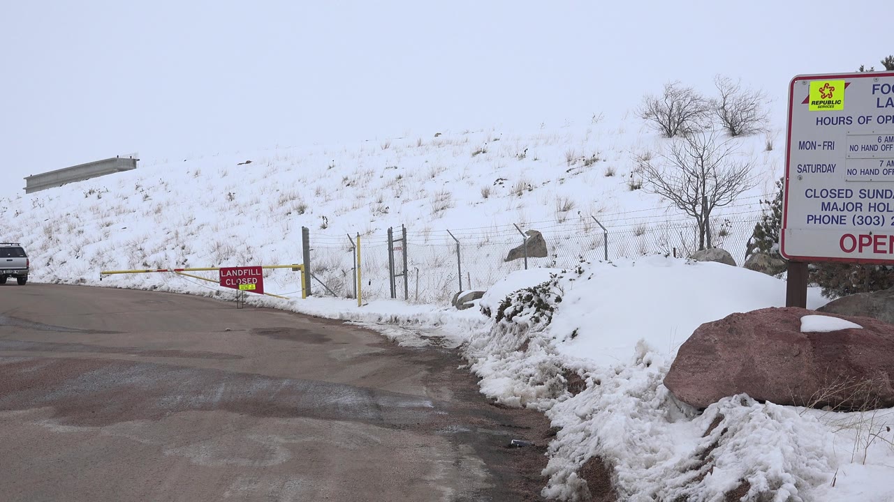 A snowy roadside entrance in Arvada, Colorado shows a wet road, red information sign, yellow gate, parked vehicle, snowbank, and no visible people.