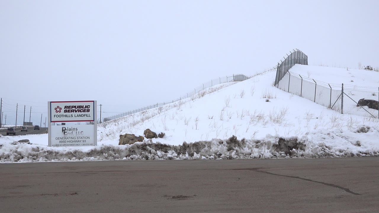 Arvada, Colorado winter roadside view with the landfill sign left of center, a fenced snow-covered ridge, exposed rocks, and no visible people.