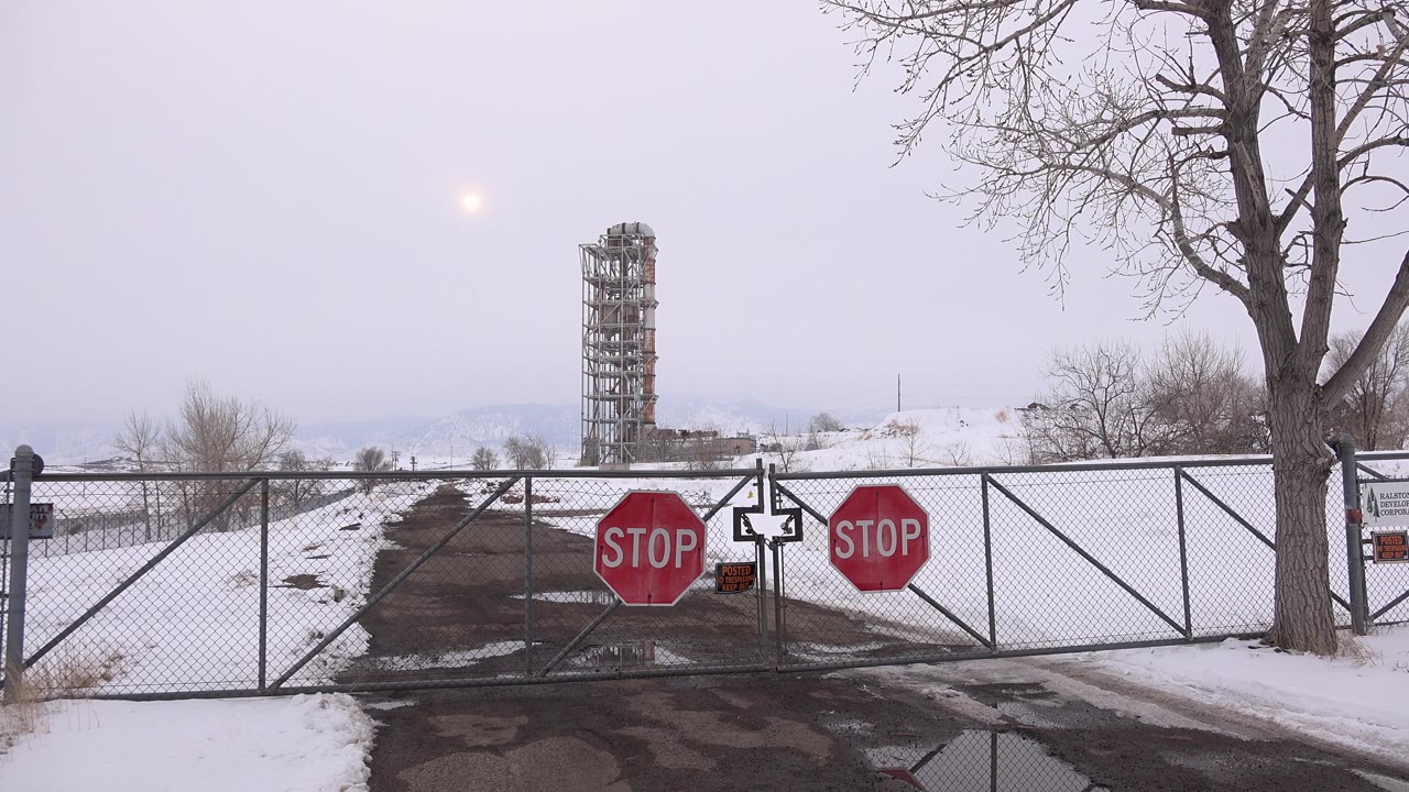 At the start of the Arvada, Colorado clip, a chain-link gate with two red stop signs blocks a snow-lined paved drive, with a leafless tree at right and no visible people.