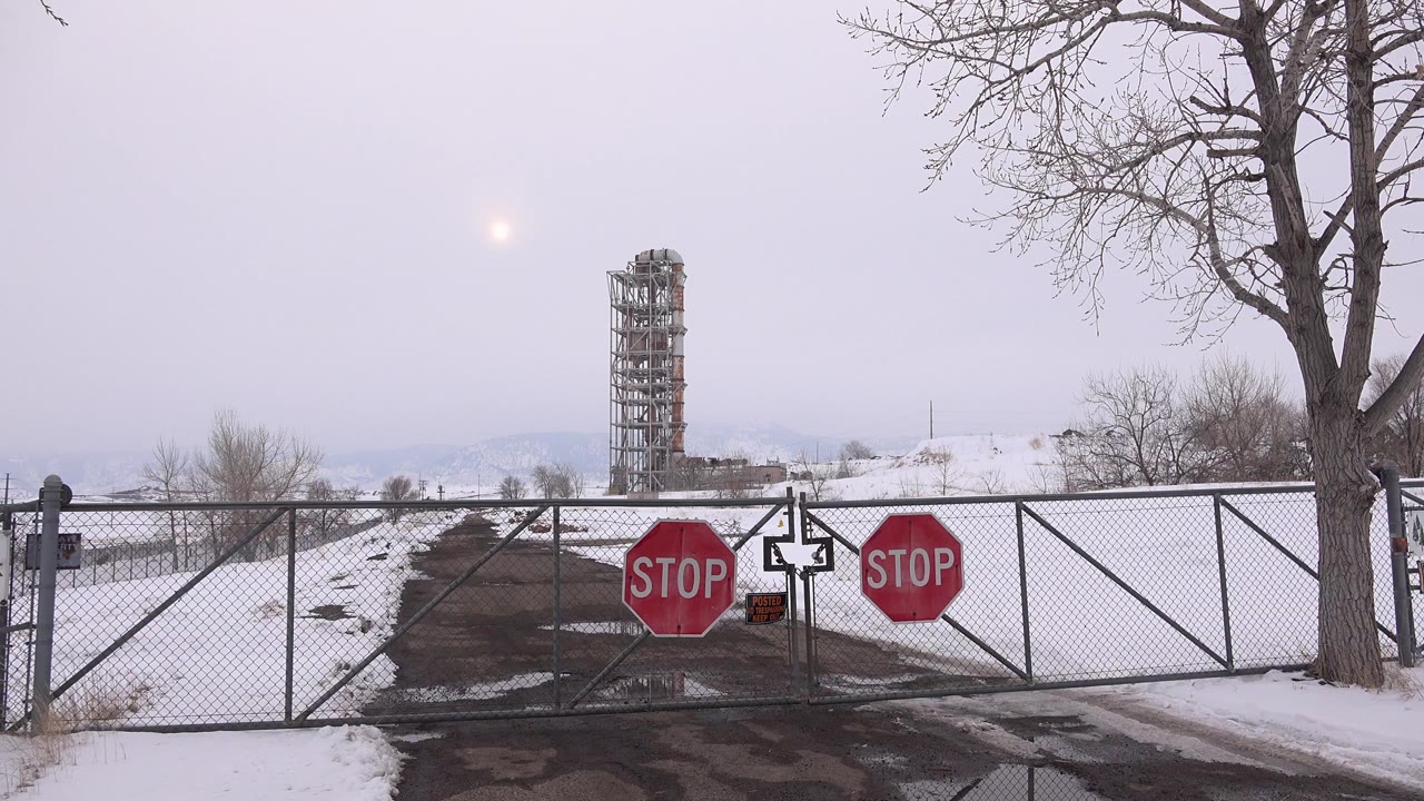 At 0:00, a closed chain-link gate with two red stop signs blocks a snowy road in Arvada, Colorado, with a bare tree at right, a metal tower in the distance, and no visible people.