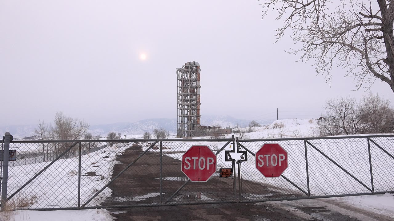 At 0:00 in Arvada, Colorado, two red stop signs on a chain-link gate block a snow-lined access road with a tall gray tower in the distance and no visible people.