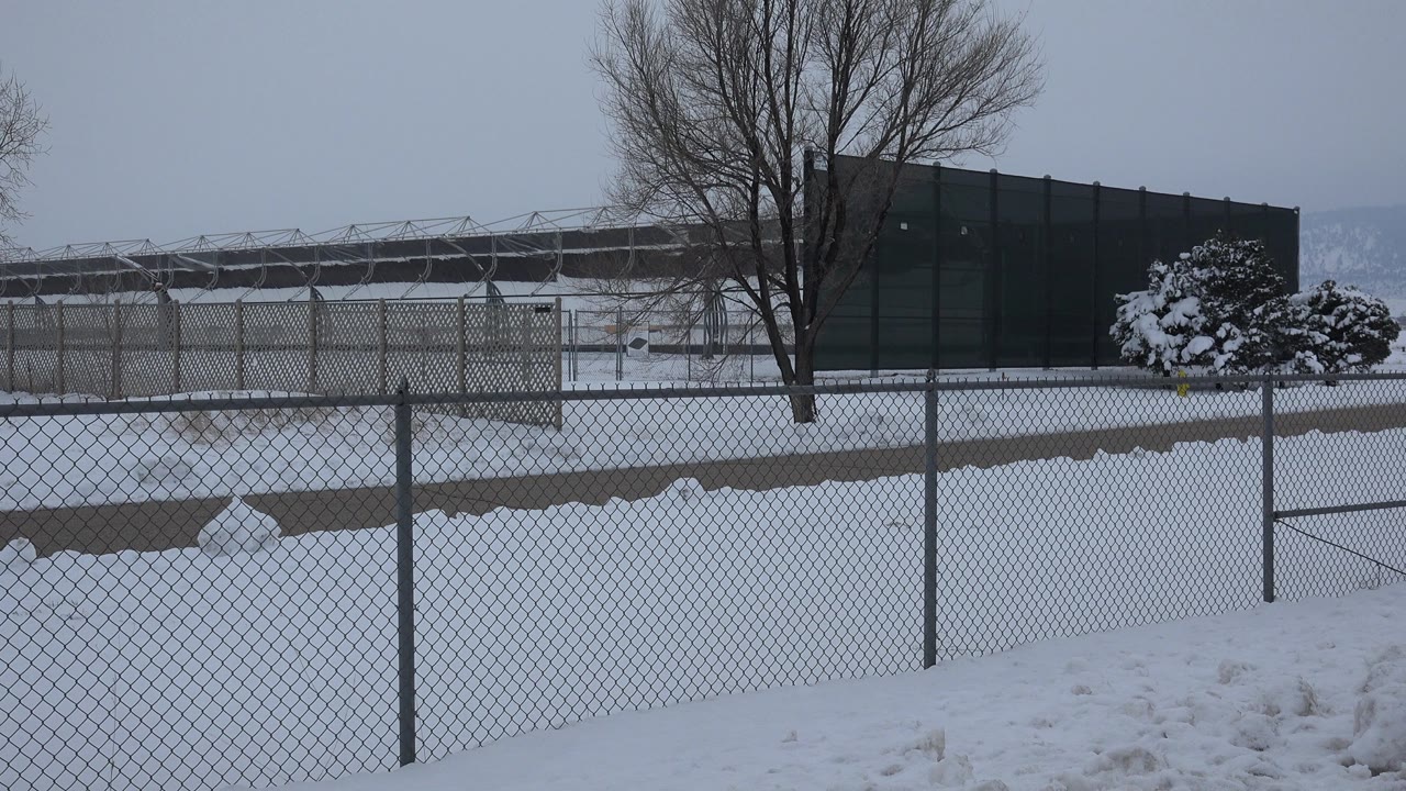 Opening view of a snow-covered fenced area in Arvada, Colorado, with chain-link fence in front, bare trees, a dark structure, and no visible people.