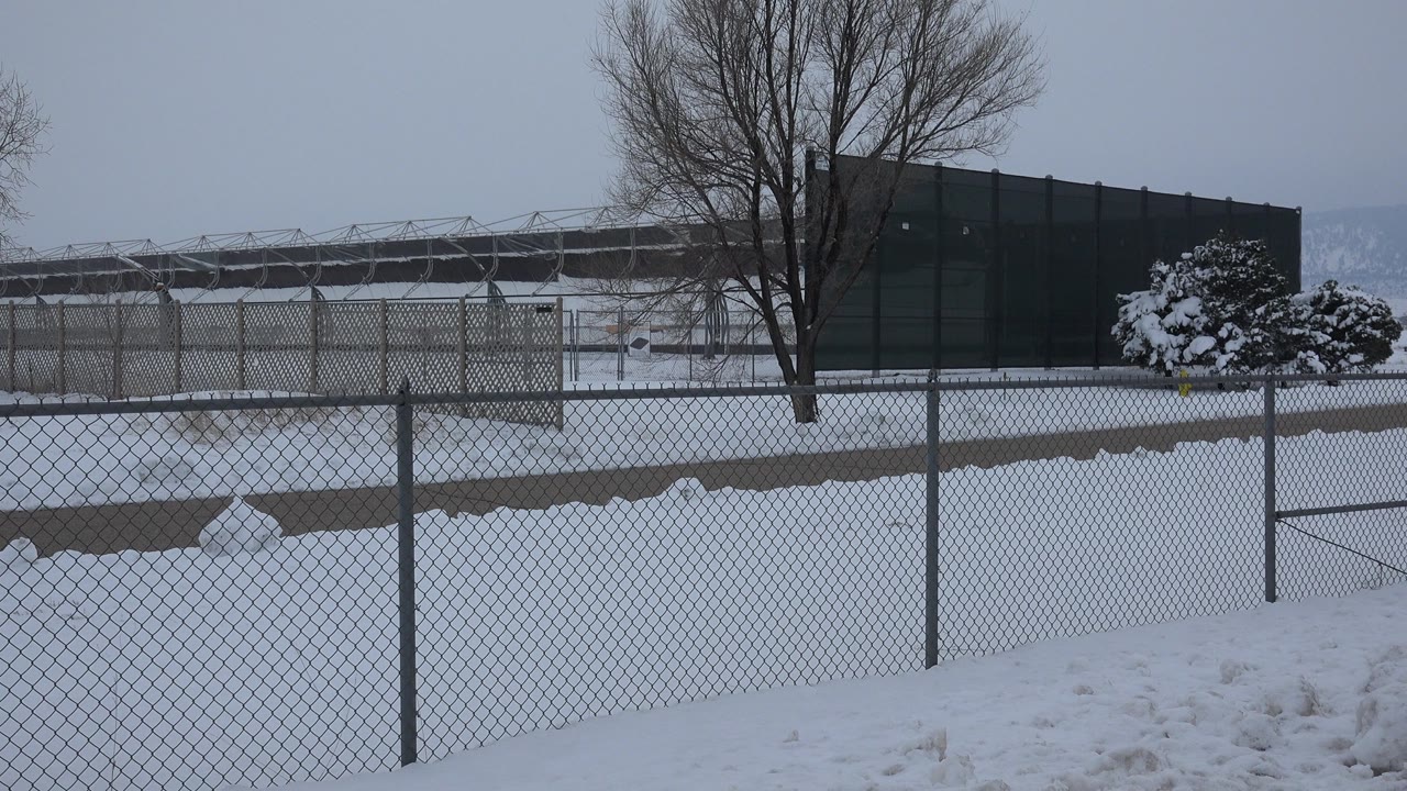 Early pan position in Arvada, Colorado, showing the fence line across the foreground, a snowy open area, bare tree trunks, a dark building, and no visible people.