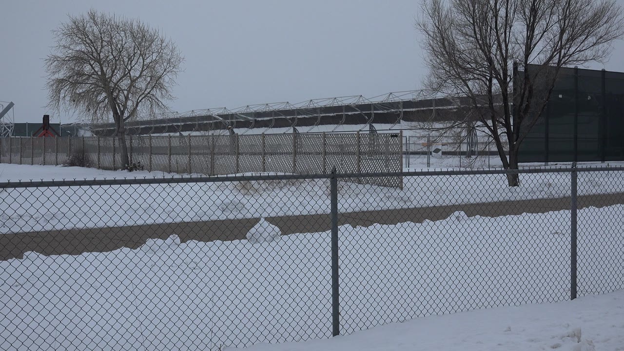 Opening frame in Arvada, Colorado showing a snowbank behind a chain-link fence, bare trees, distant industrial structures, gray sky, and no visible people.
