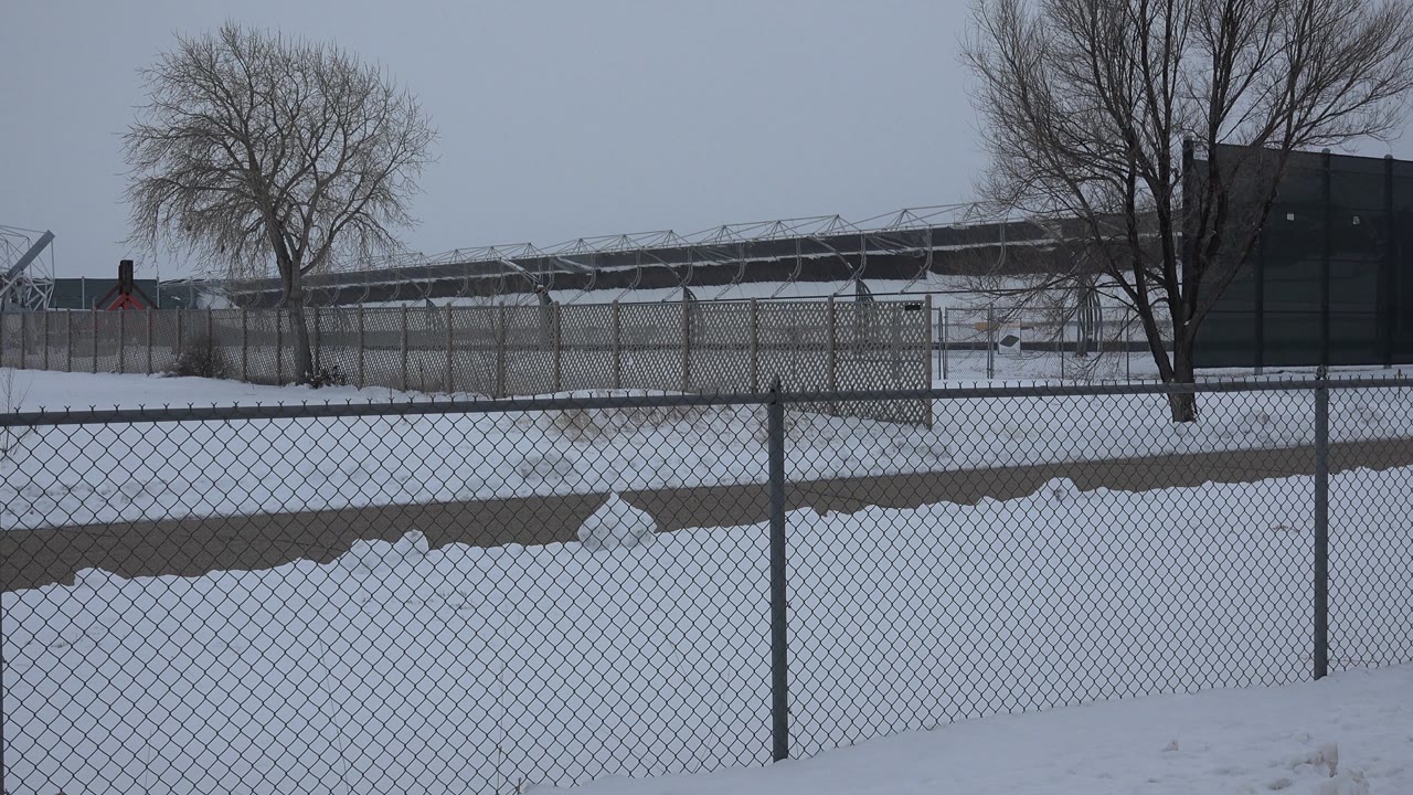 Winter fence line with snow piles, bare trees, and distant industrial roofline