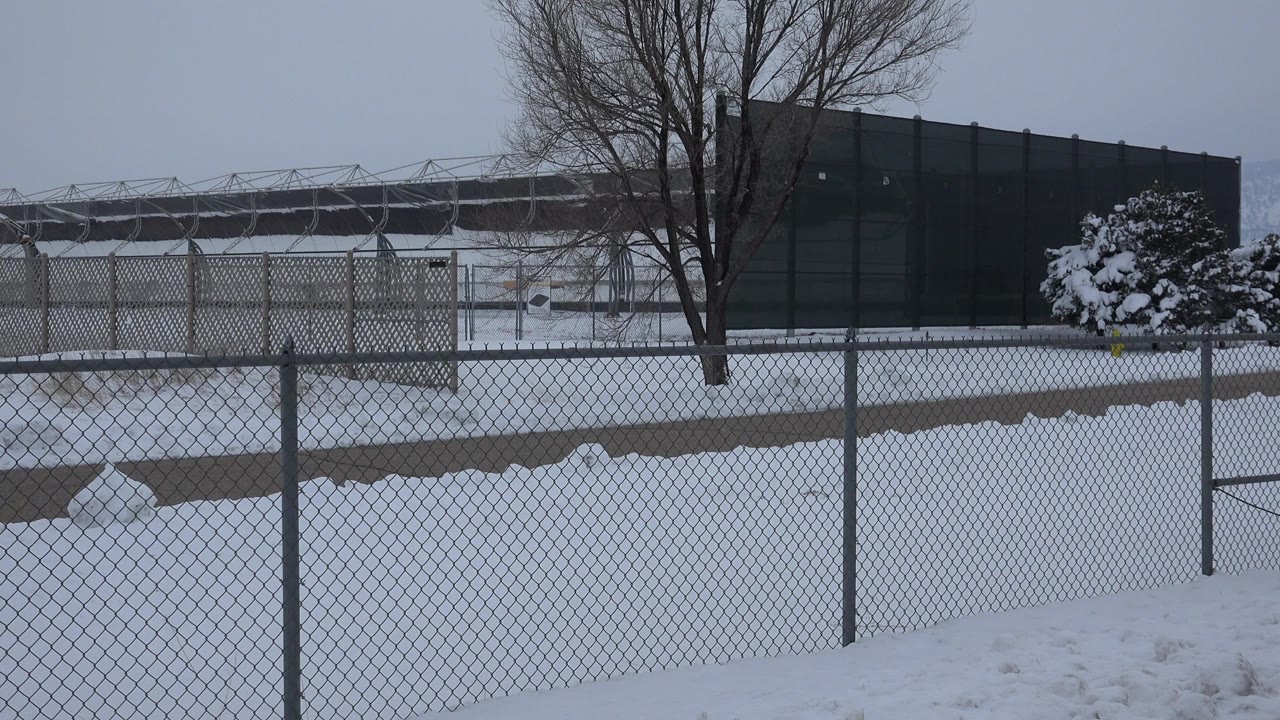 Opening frame of a snow-covered fenced area in Arvada, Colorado, with chain-link fence in front, bare trees and a dark structure behind it, and no visible people.