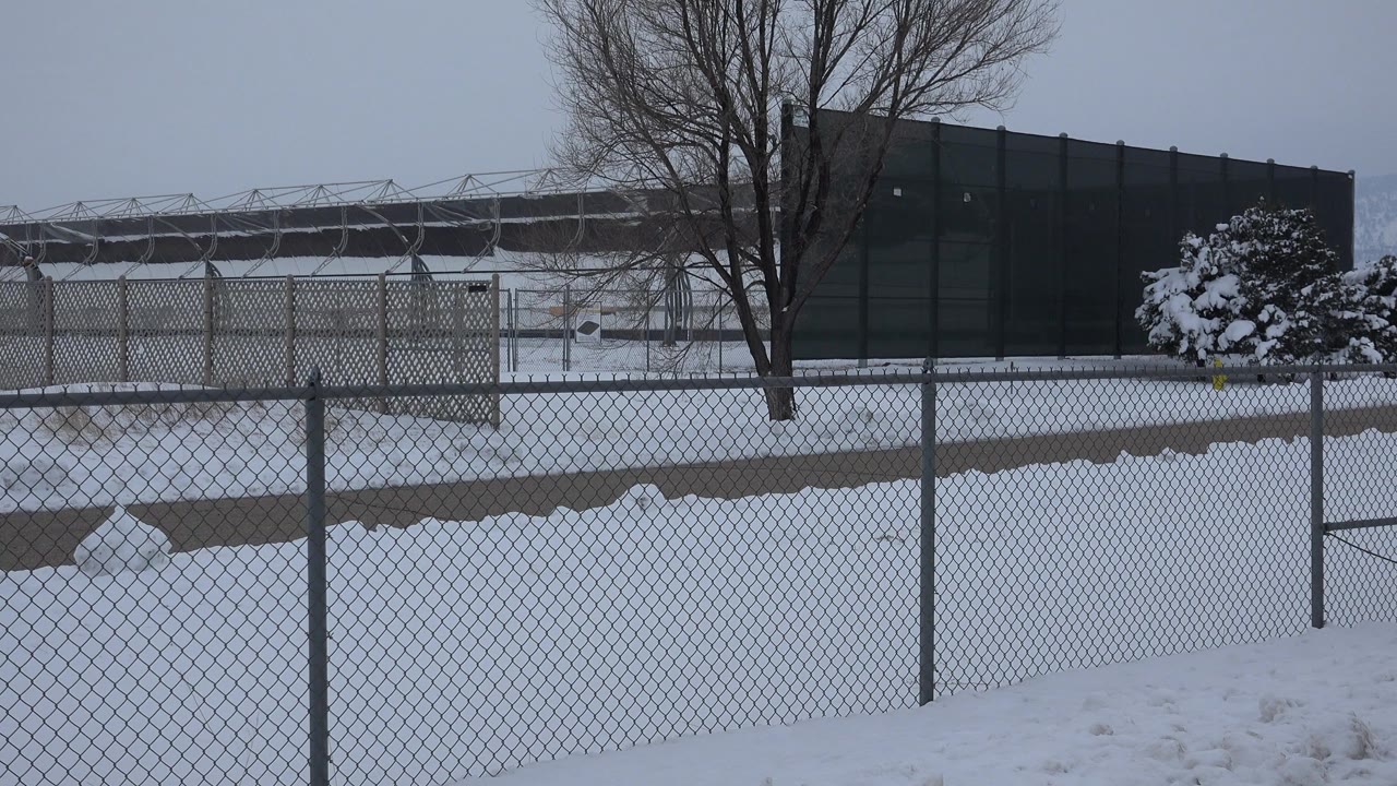 Early frame shows the Arvada winter scene with a chain-link fence crossing the foreground, snow piled across the ground, bare trees near dark panels, and no visible people.