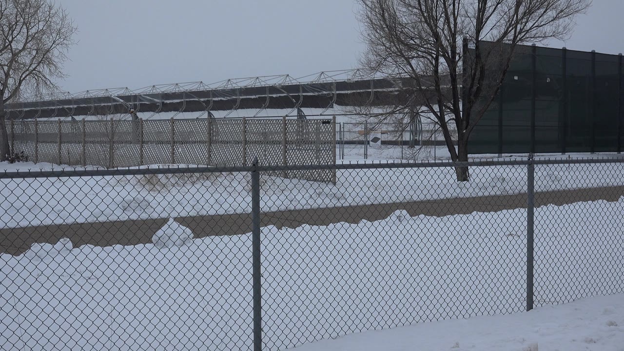 At the start of the Arvada, Colorado clip, snow covers the ground behind a chain-link fence with bare trees, a gray sky, and a dark background building, with no visible people.