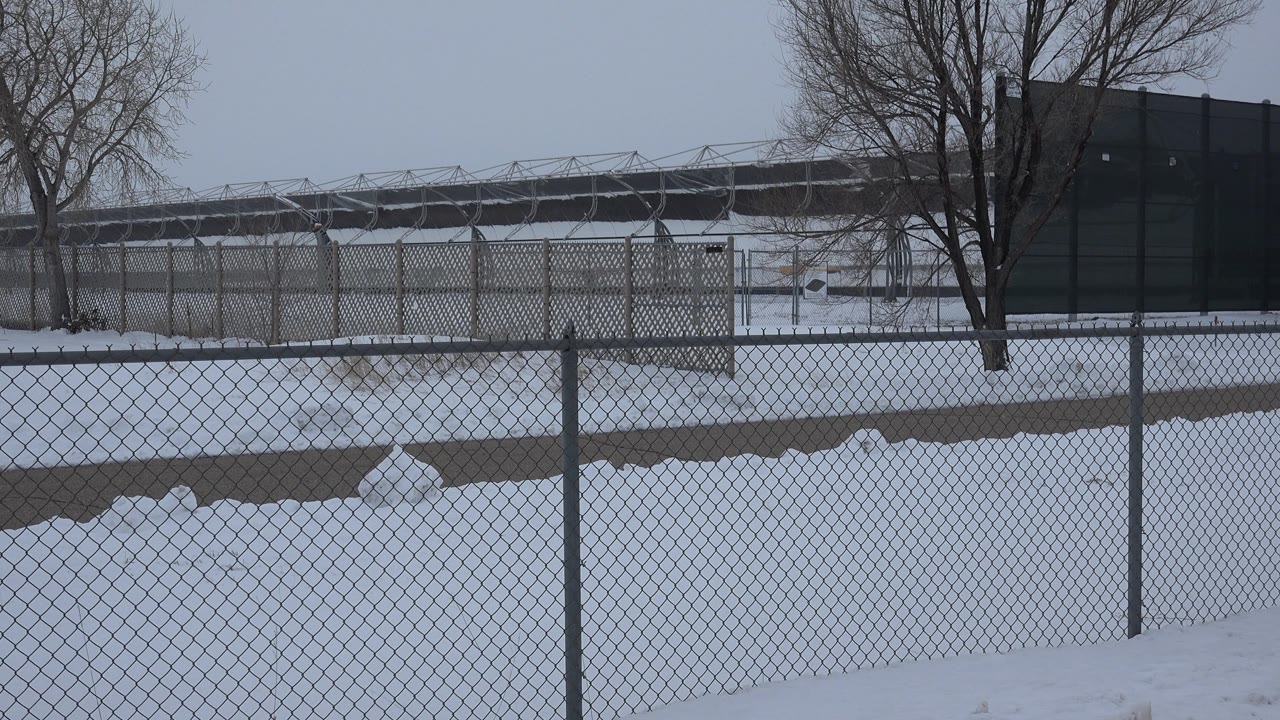 Snowy lot behind fencing with bare trees and long roofline in background.