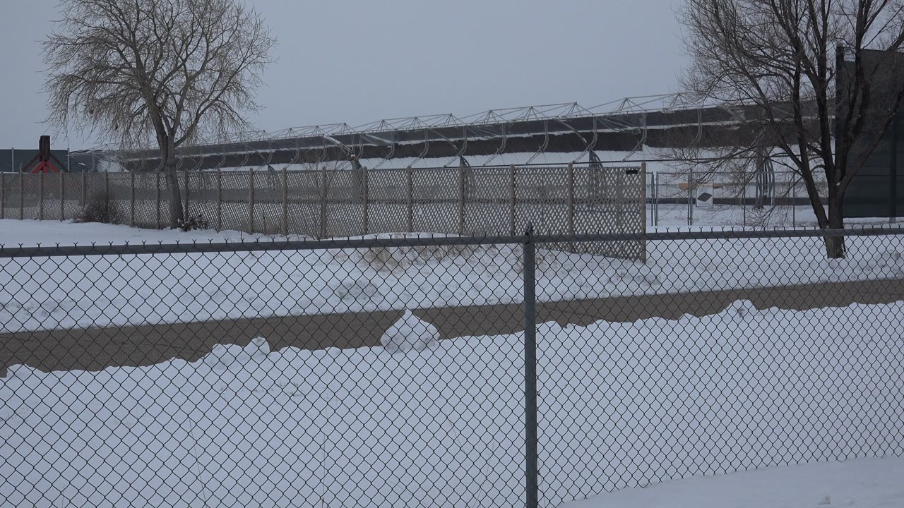 A snow-covered field in Arvada, Colorado sits behind a chain-link fence, with bare trees and a distant low structure under overcast light and no visible people.