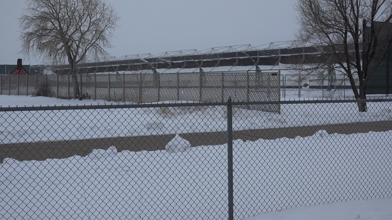 The Arvada winter view holds on fence mesh in the foreground, a snowy open area, leafless trees, and a long distant structure with no visible people.