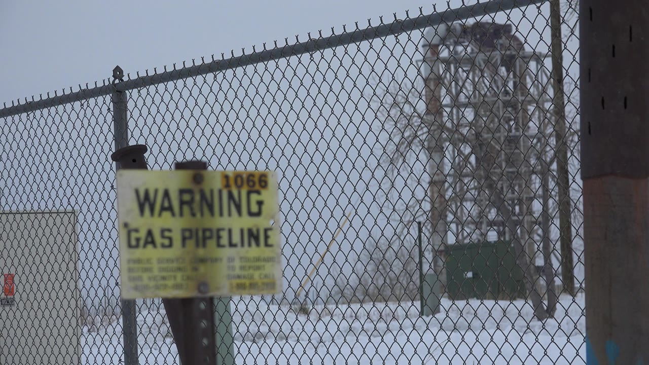 At 0:00 in Arvada, Colorado, a blurred yellow gas pipeline warning sign stands before a chain-link fence, snowy ground, and a metal utility structure, with no visible people.