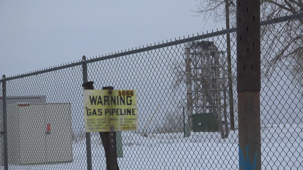 Yellow gas pipeline warning sign in front of a chain-link fence and snow-covered utility area in Arvada, Colorado, with no visible people.