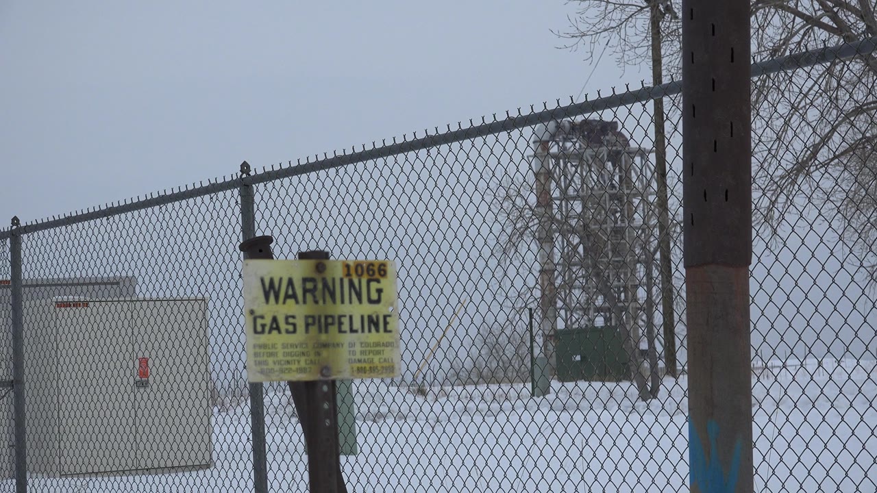 A gas pipeline warning sign stands near a dark post with chain-link fencing, utility equipment, snow, and bare trees in Arvada, Colorado, with no visible people.