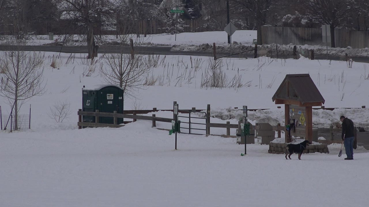 In Arvada, Colorado, a wide snowy field shows two men near a dog and a wooden kiosk, with fencing, portable toilets, roadside signs, and winter trees behind them.