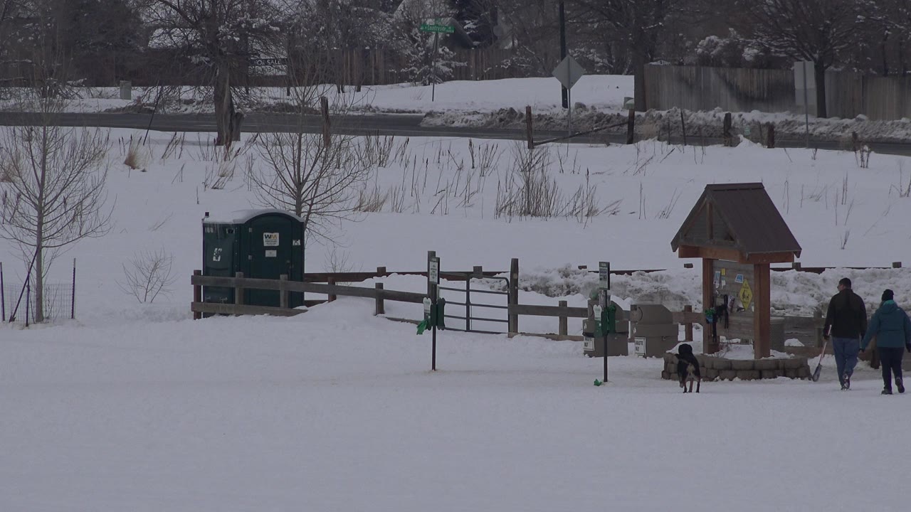 Two people and a dog walking across a snowy park area near a small roofed sign structure.