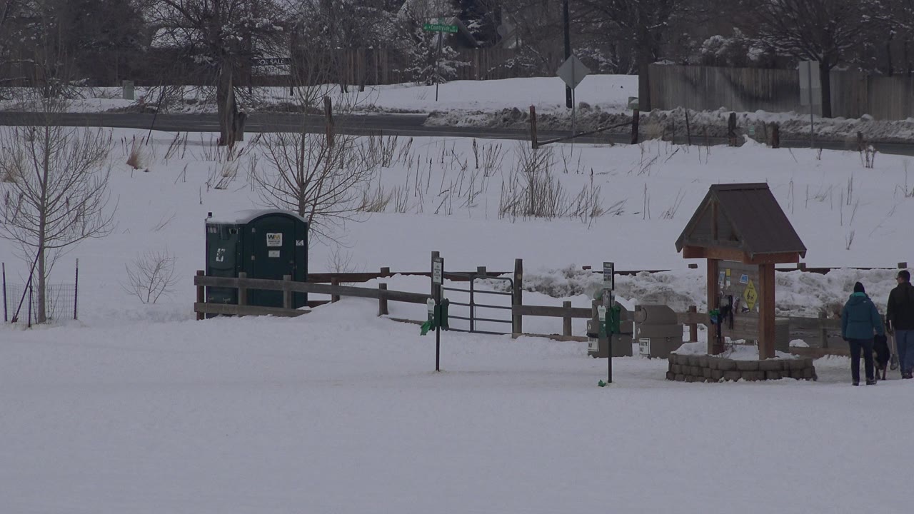 A snowy Arvada, Colorado park view shows a wooden information kiosk, fence, green portable toilets, bare trees, and two distant people near the right edge.