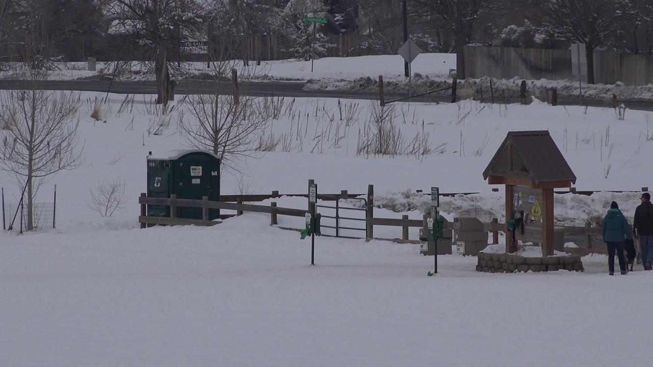 The Arvada winter park remains in a wide view as two people stand or walk beside the wooden kiosk and fence, with bare trees and snowbanks behind them.