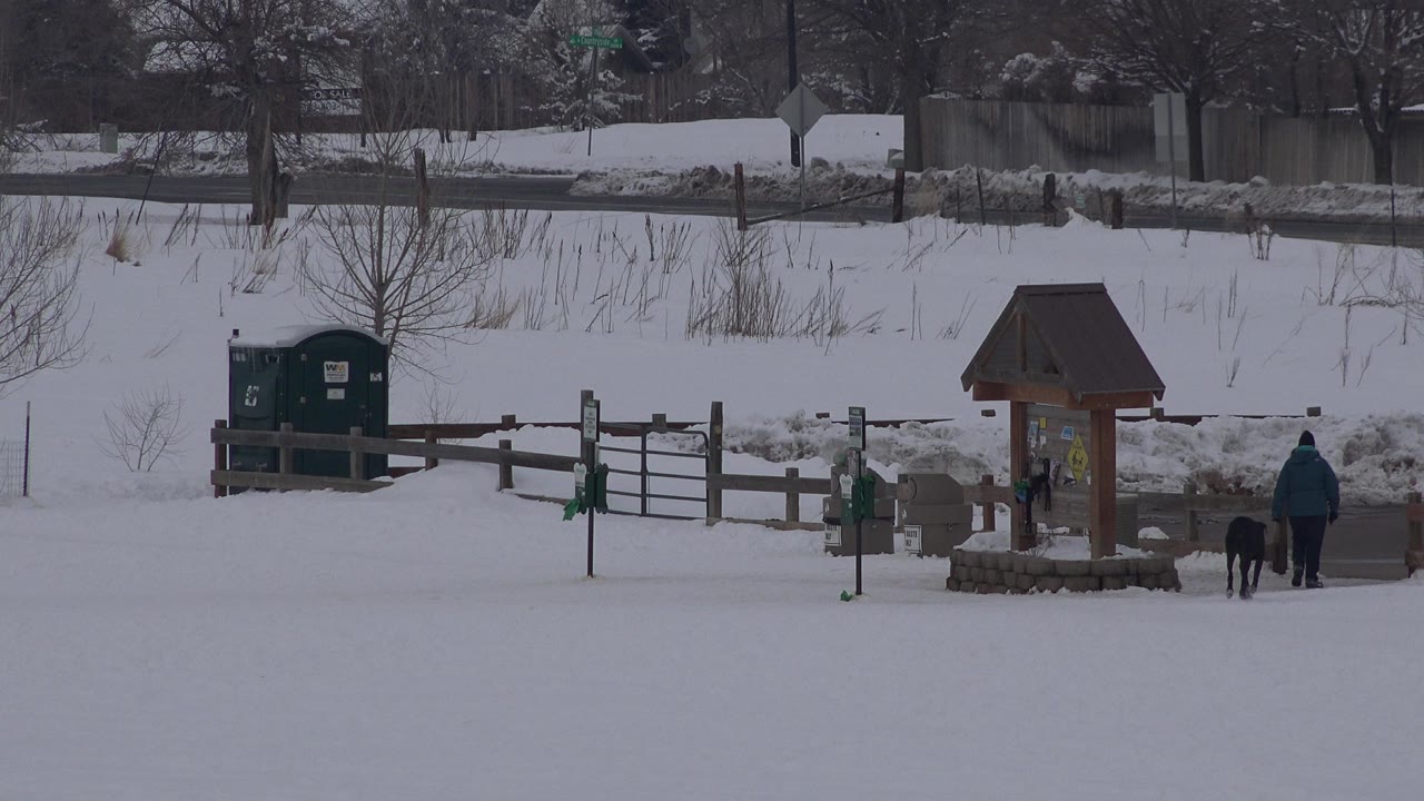 Wide Arvada, Colorado winter view with deep snow, a wooden fence, green bins, a roofed information sign, and a distant person walking a dark dog near the path.