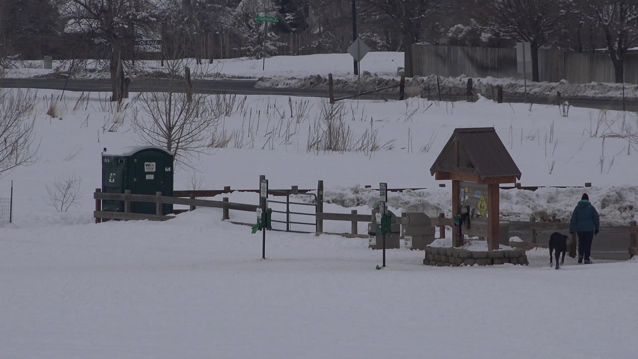 Arvada, Colorado snow scene with the roofed sign kiosk centered to the right, green bins on the left, and a distant walker with a dark dog moving along the path.