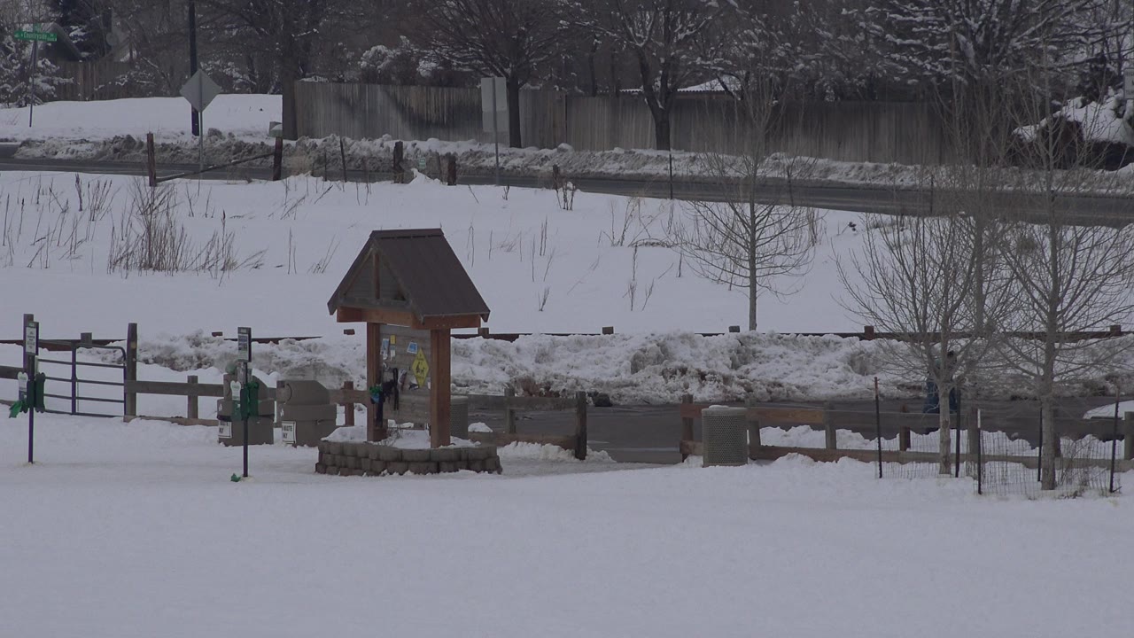 Snow-covered park with wooden kiosk, fence, road, and bare trees.