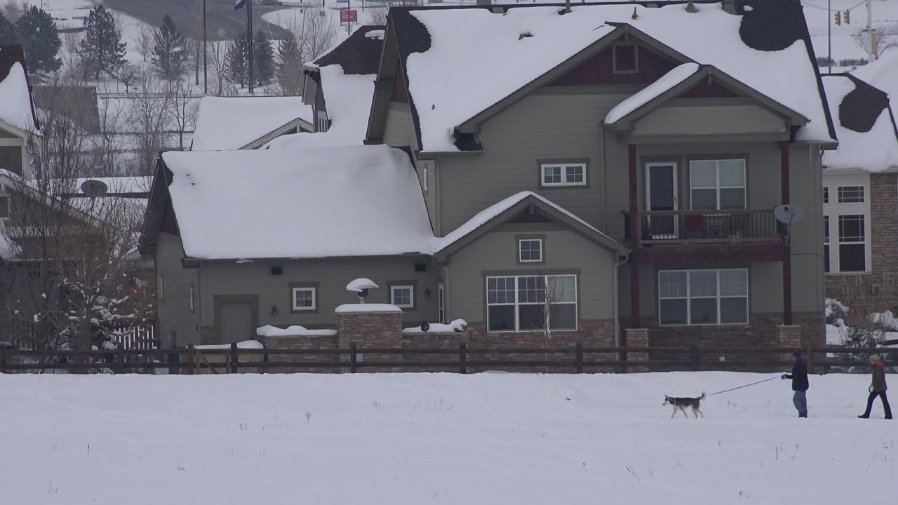 Two people walking a dog across snow in front of a brown building.