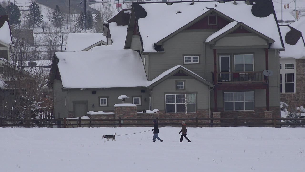 At 0:00 in Arvada, Colorado, two bundled adults and a small leashed dog move across a snowy foreground with private homes and bare trees behind them.