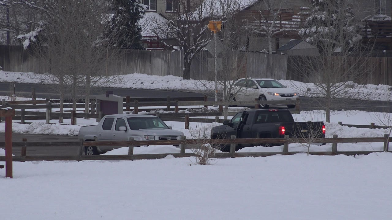 Snowy roadside with parked vehicles and houses in the background.