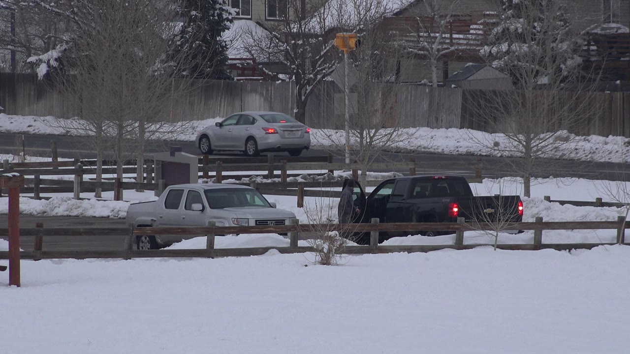Snowy suburban road with parked vehicles and a car passing in the background.