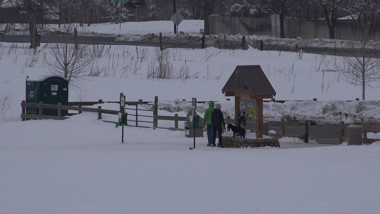 At 0:00 in Arvada, Colorado, a snowy open area shows a small roofed well or sign structure beside fencing, with distant people, a child, and a dark dog near the posts.